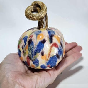 May include: A ceramic pumpkin with a cream-colored base and colorful splattered paint in blue, red, yellow, and orange. The pumpkin has a brown stem with a curled top. The pumpkin is held in a hand.