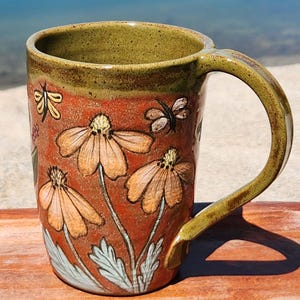 May include: A ceramic mug with a brown exterior featuring hand-painted orange flowers and butterflies. The mug has a green rim and handle. The design includes light blue leaves and a yellow center for each flower.