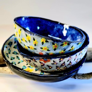 May include: A set of three small ceramic bowls with floral patterns. The bowls are stacked on top of each other, with the top bowl being blue with white and yellow flowers, the middle bowl being white with purple and orange flowers, and the bottom bowl being white with blue flowers.