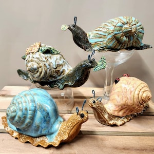 May include: Four ceramic snail sculptures in various colours and designs. The snails are arranged on a wooden surface. The snails are all different sizes and have different details, such as flowers, leaves, and spots.