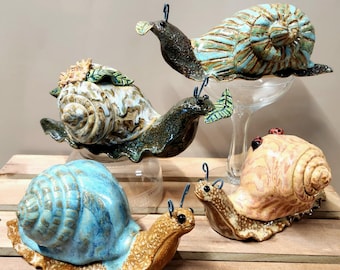 Puede incluir: Cuatro esculturas de caracoles de cerámica en varios colores y diseños. Los caracoles están dispuestos sobre una superficie de madera. Los caracoles son de diferentes tamaños y tienen diferentes detalles, como flores, hojas y manchas.