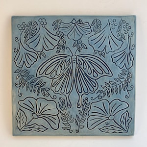 May include: A blue ceramic tile with a black butterfly design, surrounded by stylized flowers and leaves.