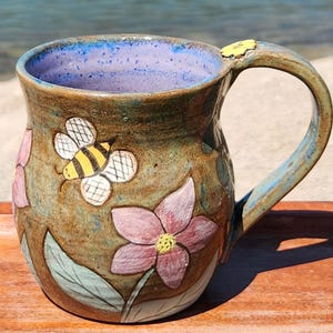 May include: A handmade ceramic mug with a brown exterior, featuring a bee and pink flower design. The interior is blue, and the handle has a small yellow flower accent. The mug is approximately 4 inches tall.