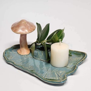 May include: A ceramic tray in a blue-green glaze with a light pink mushroom and a white candle. The tray has a leaf-like shape and a textured surface.