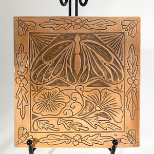 May include: A ceramic tile with a raised design of a butterfly, flowers, and leaves. The tile is a light brown color and has a dark brown glaze. The tile is on a black metal stand.