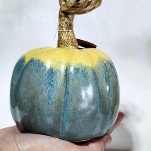 May include: A ceramic pumpkin with a yellow top and blue-green body. The stem is a twisted, textured brown with a small tag attached. The pumpkin is held in a hand, showcasing its size and shape, perfect for fall decor.
