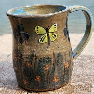 May include: A handmade ceramic mug with a brown base and a blue rim and handle. The mug is decorated with painted butterflies, orange flowers, and green foliage. The mug is designed for hot or cold beverages.