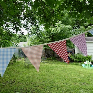 May include: A string of blue, pink, and red gingham pennant flags hanging in a backyard. The flags are strung between two trees and are decorated with a white trim.