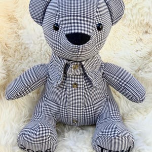 May include: A handmade teddy bear with a black and white checkered pattern. The bear is wearing a collared shirt with two buttons. The bear's paws have the text "2025" and "TBH" embroidered on them. The bear is sitting on a white, fluffy surface.