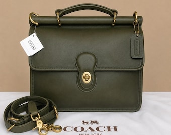 May include: An olive green leather satchel with a top handle, a turn-lock closure, and brass hardware. The bag includes a detachable shoulder strap, a hanging leather luggage tag, and a white paper tag with the Coach logo printed on the front.