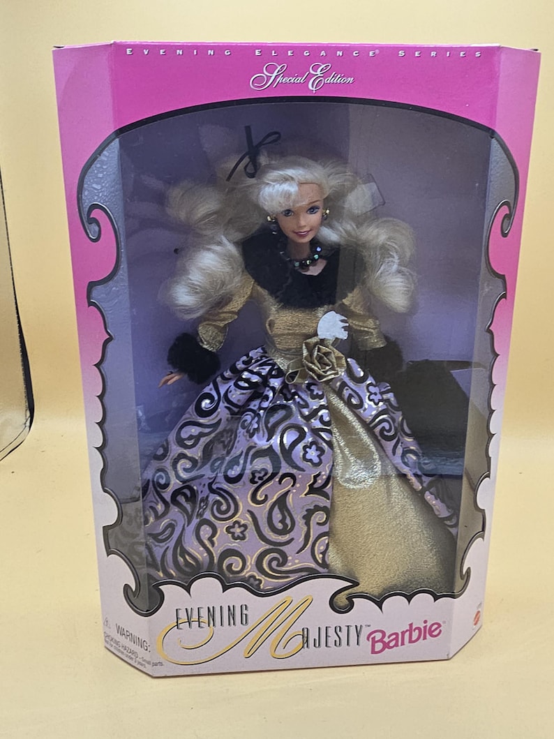 以下が含まれることがあります： Evening Elegance Series Special Edition Evening Majesty Barbieと書かれた箱に入ったブロンドの人形。人形は黒いファーのトリミングが施されたゴールドのボディスと、黒いペイズリー柄、ゴールドのバラ、黒いファーの袖口があしらわれた紫色のスカートを着用しています。