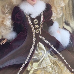 May include: A doll with long blonde hair wearing a deep burgundy velvet coat featuring white faux fur trim at the collar and cuffs, a matching fur hat, and three gold-toned decorative buttons. The coat opens to reveal a gold metallic skirt.