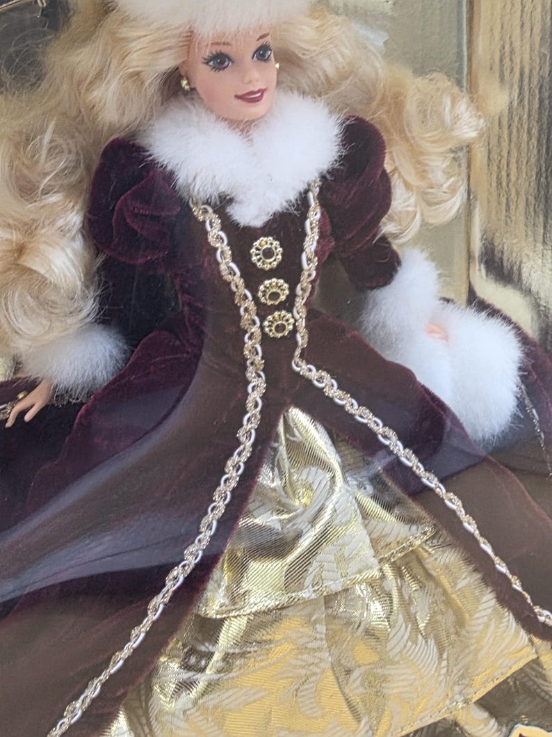 May include: A doll with long blonde hair wearing a deep burgundy velvet coat featuring white faux fur trim at the collar and cuffs, a matching fur hat, and three gold-toned decorative buttons. The coat opens to reveal a gold metallic skirt.