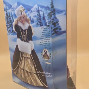 May include: A rectangular cardboard box containing a Happy Holidays Barbie doll. The packaging features a winter landscape with snow-covered mountains and trees. Text on the box includes Happy Holidays, Barbie, and a description of the doll's attire.