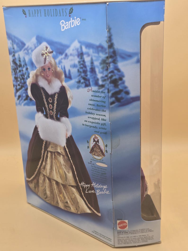 May include: A rectangular cardboard box containing a Happy Holidays Barbie doll. The packaging features a winter landscape with snow-covered mountains and trees. Text on the box includes Happy Holidays, Barbie, and a description of the doll's attire.