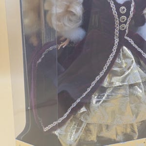 May include: A doll with blonde hair wearing a deep burgundy velvet gown with a white faux fur collar and cuffs. The dress features gold metallic trim and a layered gold skirt. The doll is inside a clear plastic box labeled Special Edition.