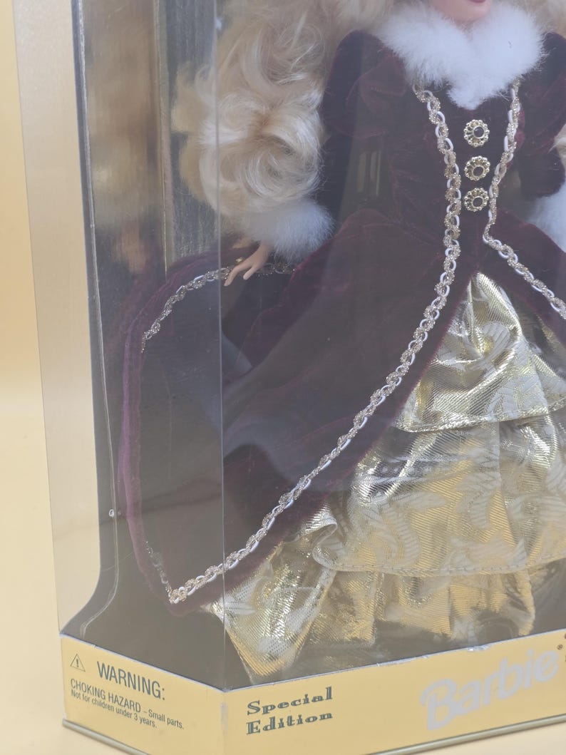 May include: A doll with blonde hair wearing a deep burgundy velvet gown with a white faux fur collar and cuffs. The dress features gold metallic trim and a layered gold skirt. The doll is inside a clear plastic box labeled Special Edition.