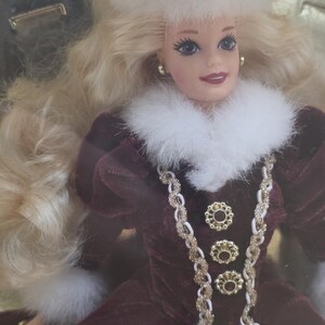 May include: A doll with long blonde hair wearing a white fur hat and a dark maroon velvet dress. The dress features a white fur collar, gold-toned braided trim, and three circular gold buttons down the front. The doll has gold-toned stud earrings.