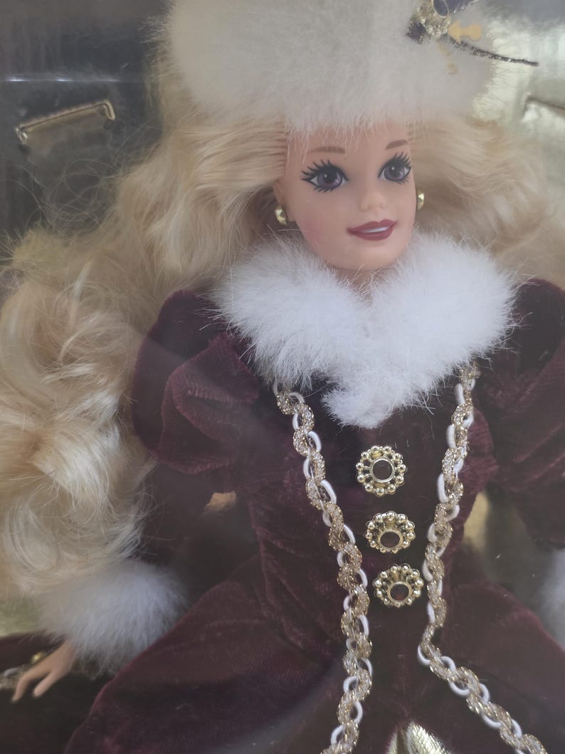May include: A doll with long blonde hair wearing a white fur hat and a dark maroon velvet dress. The dress features a white fur collar, gold-toned braided trim, and three circular gold buttons down the front. The doll has gold-toned stud earrings.