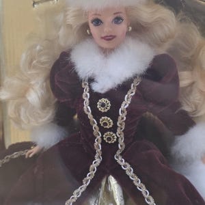 May include: A blonde doll wearing a deep burgundy velvet coat with white faux fur trim at the collar and cuffs. The coat features three gold-toned decorative buttons and braided trim. The doll wears a matching white faux fur hat with a gold accent.
