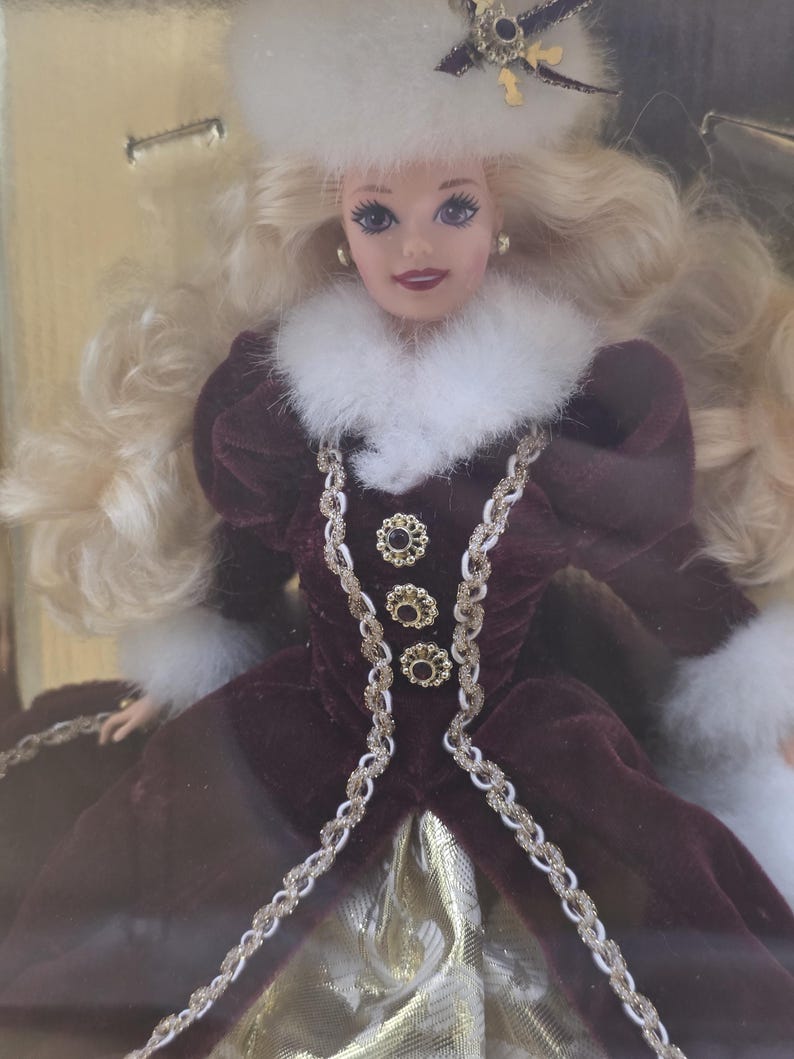 May include: A blonde doll wearing a deep burgundy velvet coat with white faux fur trim at the collar and cuffs. The coat features three gold-toned decorative buttons and braided trim. The doll wears a matching white faux fur hat with a gold accent.