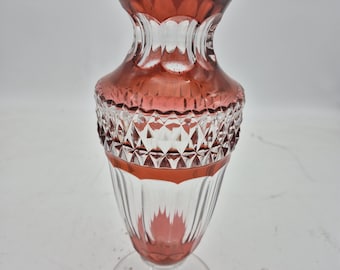 Puede incluir: Un jarrón de cristal transparente con borde y detalles en rojo rubí. El jarrón tiene una parte superior acampanada y un patrón tallado en diamante en el centro. La base es un pie claro y circular.