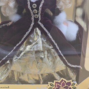 May include: A doll with blonde hair wearing a deep burgundy velvet gown with white faux fur trim at the collar and cuffs. The dress features gold metallic fabric layers, decorative gold buttons, and braided trim. The packaging displays the text Barbie.