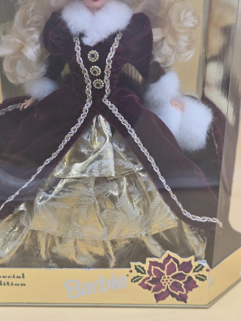 May include: A doll with blonde hair wearing a deep burgundy velvet gown with white faux fur trim at the collar and cuffs. The dress features gold metallic fabric layers, decorative gold buttons, and braided trim. The packaging displays the text Barbie.