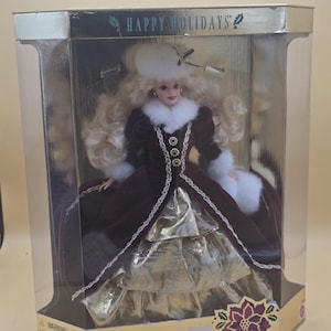 以下が含まれることがあります： Happy Holidays Special Editionのバービー人形がディスプレイボックスに入っています。人形は長いブロンドの髪、白いファーの帽子、白いファーのトリミングが施されたダークバーガンディのベルベットのコートを着用しています。下にはゴールドのメタリックなスカートが見えます。箱にはゴールドのアクセントが施されています。