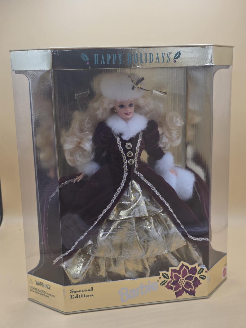 May include: A Happy Holidays Special Edition Barbie doll in a display box. The doll has long blonde hair, a white fur hat, and a dark burgundy velvet coat with white fur trim. Underneath, a gold metallic skirt is visible. The box features gold accents.