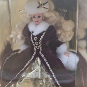 May include: A blonde doll in a box labeled HAPPY HOLIDAYS, wearing a deep burgundy velvet gown with white faux fur trim, a matching white fur hat with a dark star-shaped ornament, and a gold metallic skirt with a textured pattern.