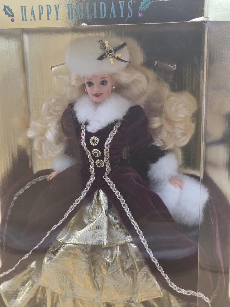 May include: A blonde doll in a box labeled HAPPY HOLIDAYS, wearing a deep burgundy velvet gown with white faux fur trim, a matching white fur hat with a dark star-shaped ornament, and a gold metallic skirt with a textured pattern.