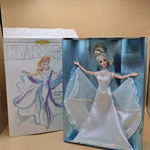 以下が含まれることがあります： ClassiqueコレクションのStarlight Barbie人形。オリジナルパッケージ入り。人形は、きらめくシアーなケープとシルバーのアクセントが付いた白いサテンのガウンを着用しています。箱には人形のイラストとClassiqueという文字が印刷されています。