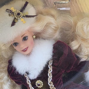 May include: A doll with long blonde hair wearing a white faux fur hat with a gold and dark ribbon accent, and a deep burgundy velvet coat featuring a white faux fur collar, gold buttons with dark centers, and a braided gold and white trim.