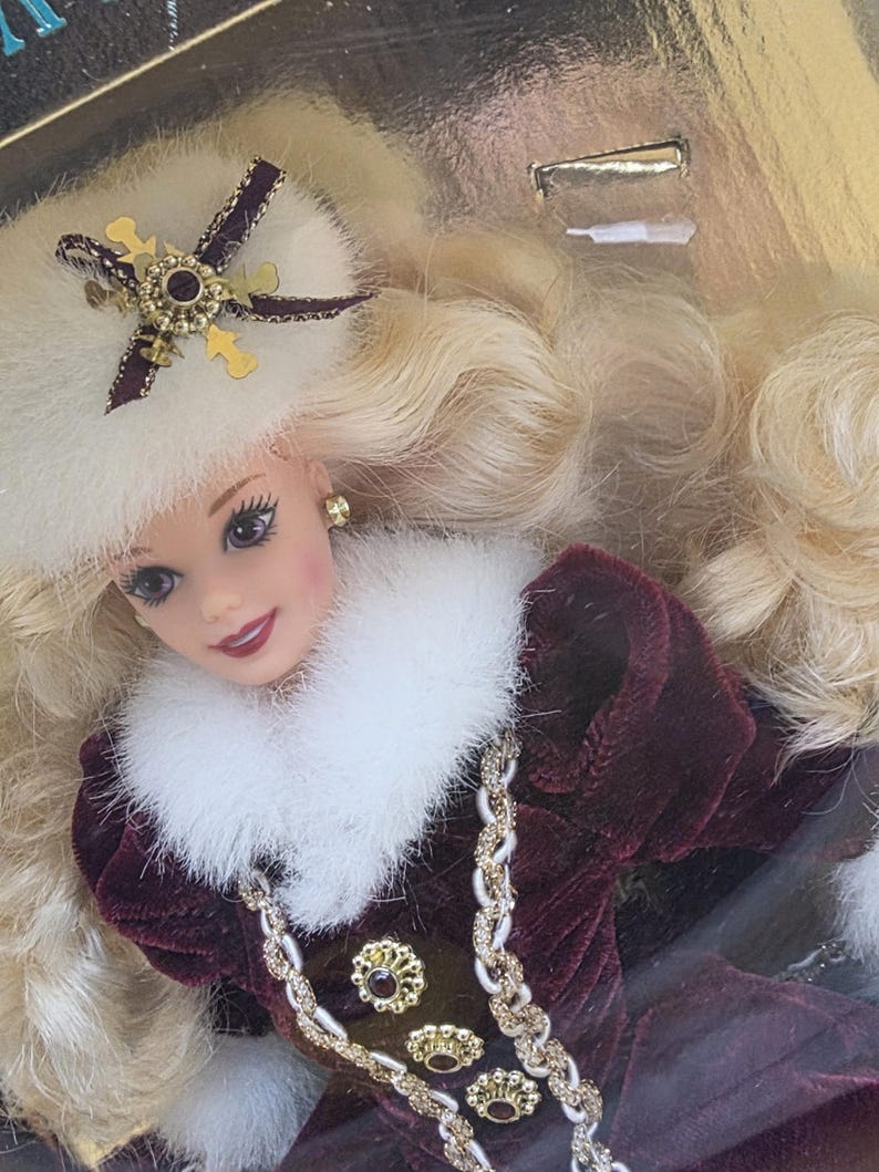 May include: A doll with long blonde hair wearing a white faux fur hat with a gold and dark ribbon accent, and a deep burgundy velvet coat featuring a white faux fur collar, gold buttons with dark centers, and a braided gold and white trim.