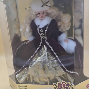 May include: A Special Edition Barbie doll in a display box labeled Happy Holidays. The doll has blonde hair, a white fur hat with a star ornament, and a deep burgundy velvet coat with white fur trim. The dress features a gold metallic skirt.