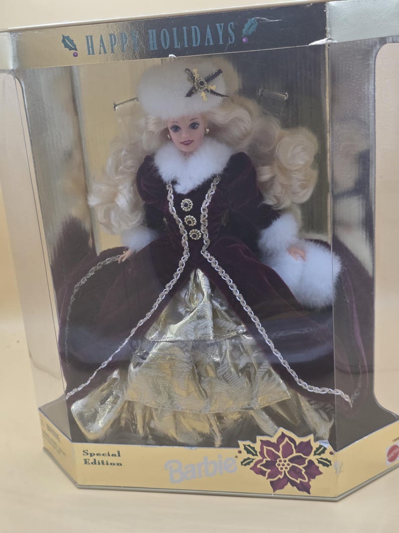 May include: A Special Edition Barbie doll in a display box labeled Happy Holidays. The doll has blonde hair, a white fur hat with a star ornament, and a deep burgundy velvet coat with white fur trim. The dress features a gold metallic skirt.