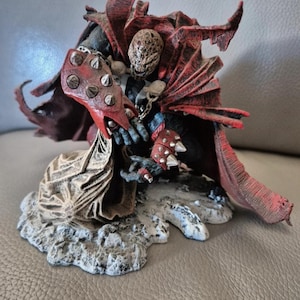 Op de afbeelding: Een rood en zwart action figure van Spawn, een stripfiguur, in een knielende houding. De figuur houdt een ketting vast in zijn rechterhand en heeft een rode cape met een gescheurde rand. De figuur staat op een rotsachtige basis.
