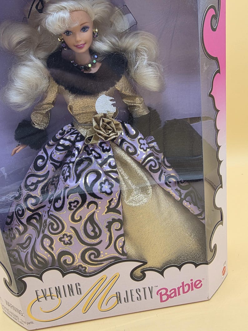 以下が含まれることがあります： Evening Majesty Barbieと書かれた箱に入ったブロンドのバービー人形。人形はダークブラウンのフェイクファーの襟が付いたゴールドのボディスと、パープルとブラックのペイズリー柄のロングスカートを着用しており、ウエストにはゴールドのバラのアクセントが付いています。