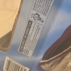 May include: The back of a Happy Holidays Barbie doll packaging featuring consumer information text, a Mattel logo, and a barcode. The visible portion of the doll shows a burgundy gown with faux fur trim and a golden brocade underskirt.