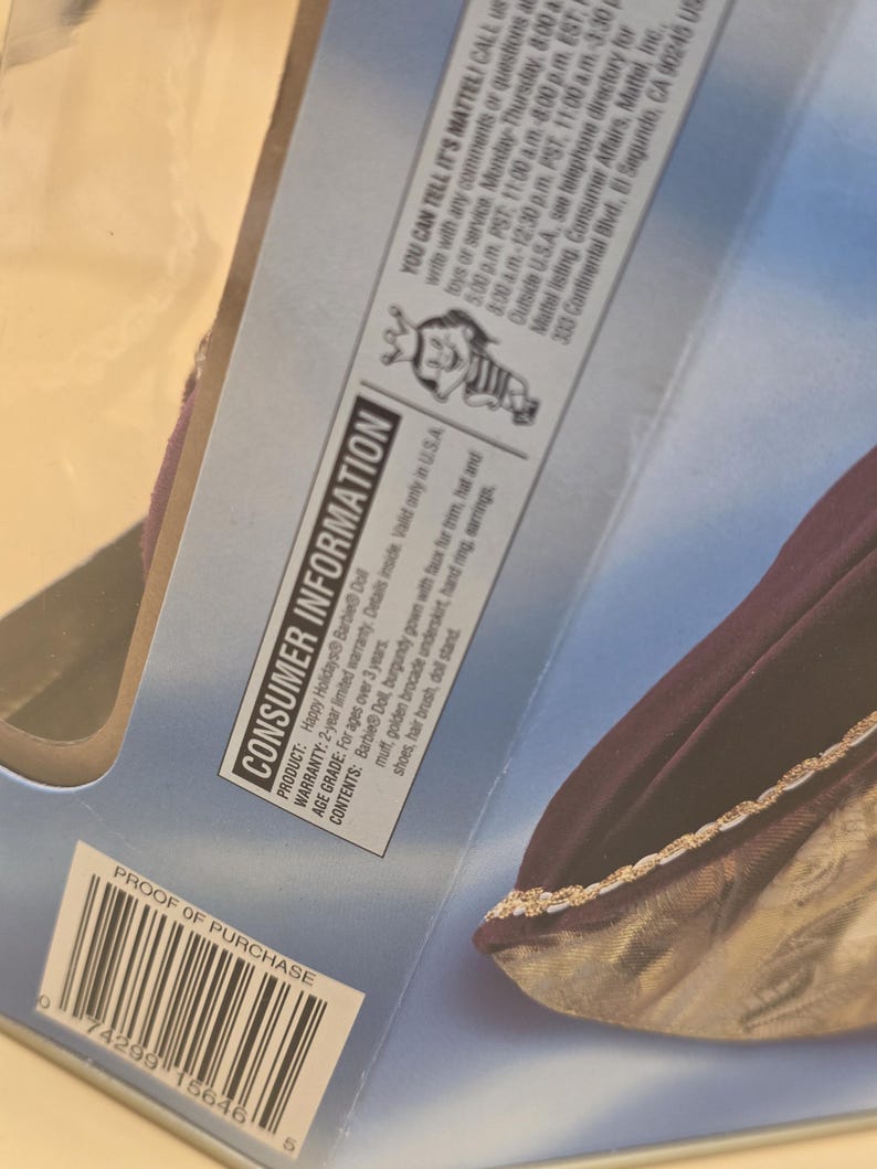 May include: The back of a Happy Holidays Barbie doll packaging featuring consumer information text, a Mattel logo, and a barcode. The visible portion of the doll shows a burgundy gown with faux fur trim and a golden brocade underskirt.