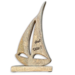 Könnte beinhalten: Holzsegelschiff-Skulptur mit natürlichem Finish. Das Segel hat ein ausgeschnittenes Design und die Worte "Ahoi Udo!" in Schwarz. Die Skulptur ist auf einem rechteckigen Sockel montiert, was ein nautisches Dekorationsobjekt ergibt.