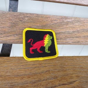 Puede incluir: Un parche bordado negro, amarillo, rojo y verde con una silueta de león. El león está mirando hacia la derecha y tiene un diseño de rayo en su espalda.