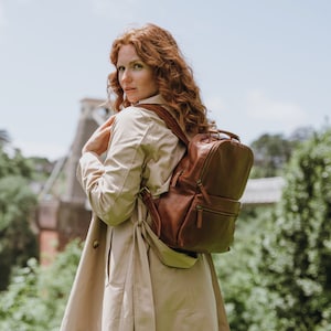 May include: A woman wearing a beige trench coat carries a brown leather backpack with multiple pockets and zips.