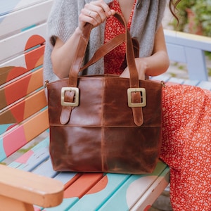 May include: A brown leather tote bag featuring two vertical straps on the front, each secured with a large, square, brushed gold-tone metal buckle. The bag has a smooth, polished finish with visible stitching along the center seams and top edge.