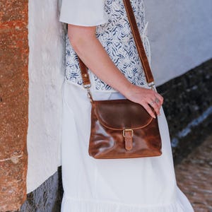 May include: A brown leather crossbody bag with a flap closure and a buckle. The bag has a long adjustable strap and is worn over the shoulder. The leather has a distressed look. The bag is a medium size.