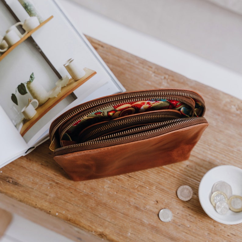 K&ouml;nnte beinhalten: Eine braune Leder-Rei&szlig;verschlusstasche mit einem bunten Blumenfutter. Die Tasche ist offen und der Rei&szlig;verschluss ist sichtbar. Die Tasche liegt auf einer Holzoberfl&auml;che.