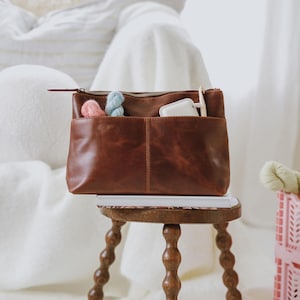 May include: A brown leather pouch with a top zipper closure and two exterior front pockets. The pouch contains a small pink yarn skein, a light blue yarn skein, and a white crochet hook. The pouch rests on a white book atop a wooden stool.