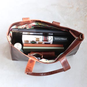 Open brown leather tote bag filled with books, a laptop, and a water bottle. The bag has a floral patterned lining and a zipper closure.