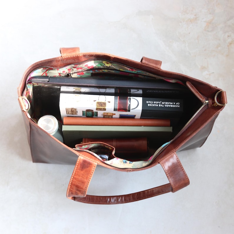 Open brown leather tote bag filled with books, a laptop, and a water bottle. The bag has a floral patterned lining and a zipper closure.